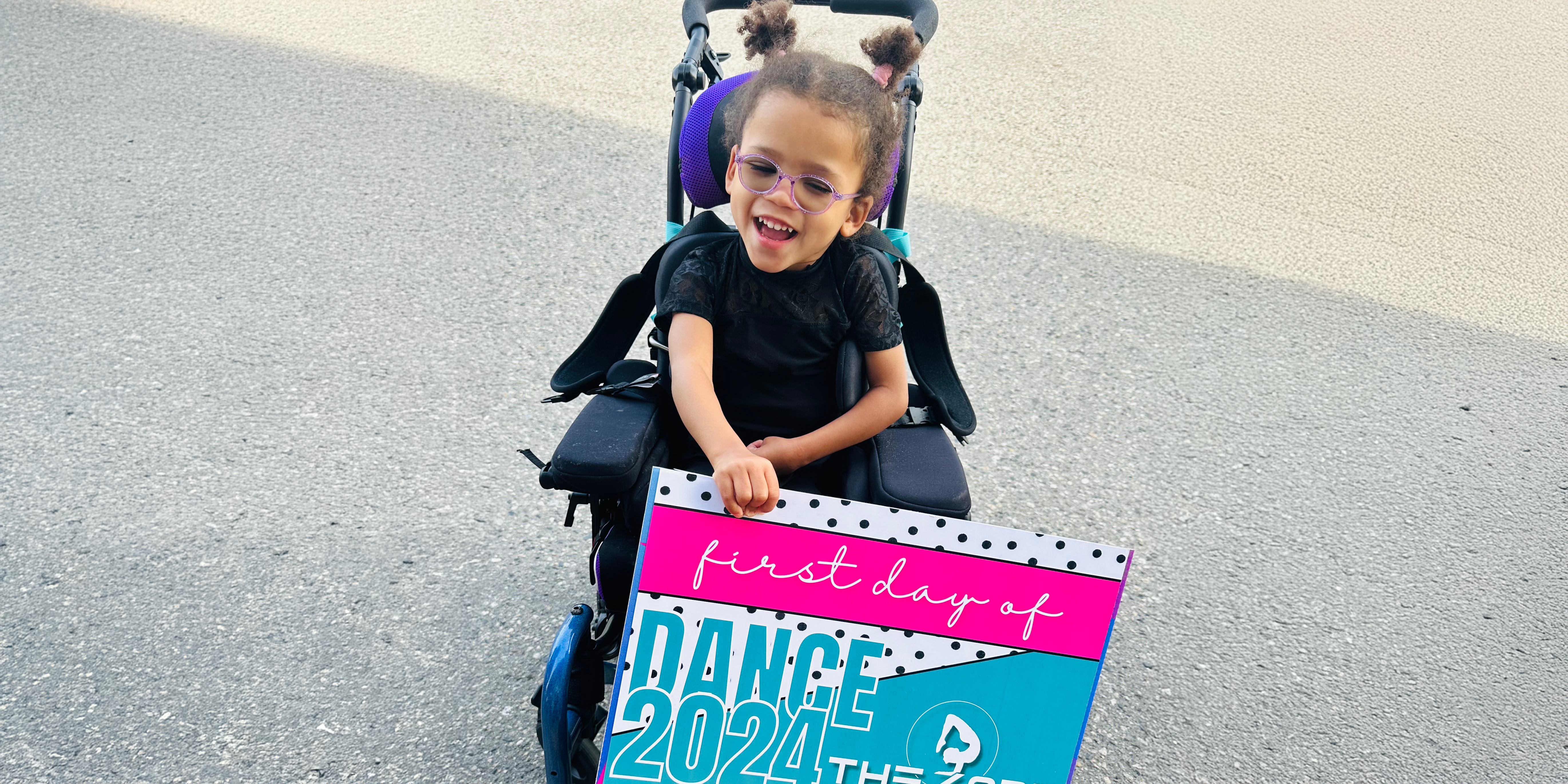 Prescott smiles from her wheelchair, holding a sign saying 'first day of dance.' Having her regular physiotherapy sessions covered by Variety BC gives her family the financial margin to enroll her in activities she loves, like dance class.