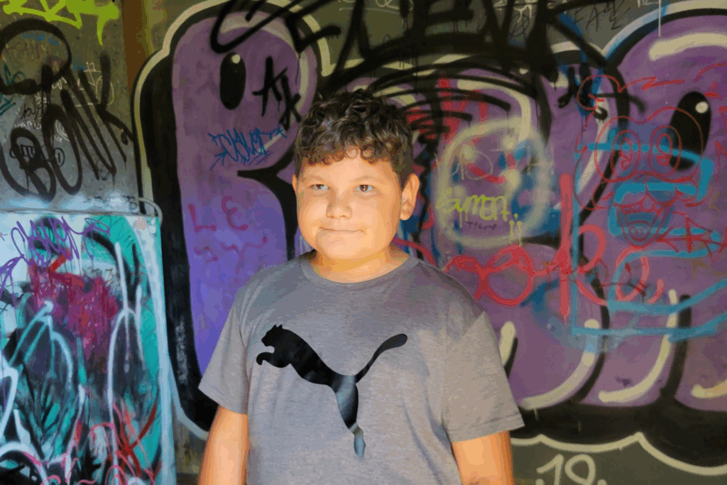 A young boy with curly hair gives a slight smile at the camera, posing in front of wall covered in graffiti.
