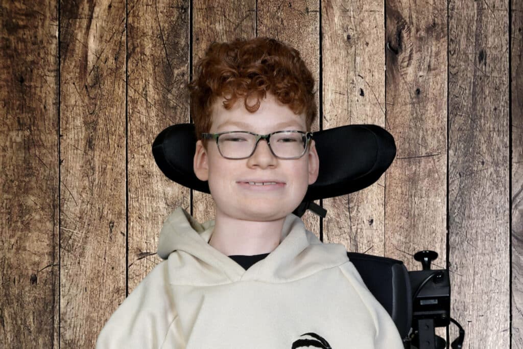 Nicolas, a young boy with curly red hair and glasses, poses in his power wheelchair against a wooden backdrop.